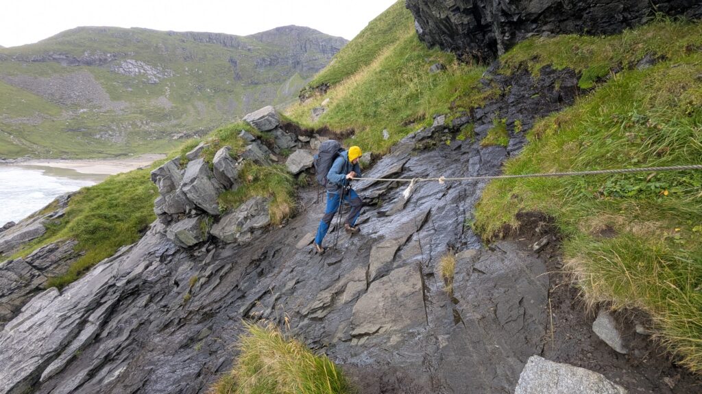 Hiker crossing an exposed rope-assisted section of the trail between Kvalvika and Vestervika beaches
