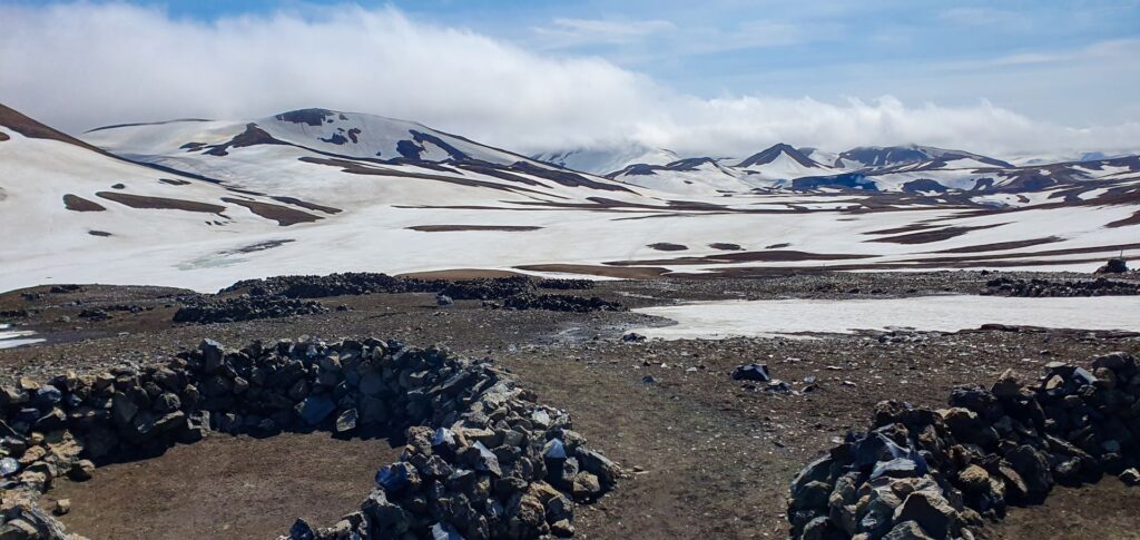 Stone wind shelters at Hrafntinnusker Campsite with snow-covered mountains in the Icelandic Highlands.